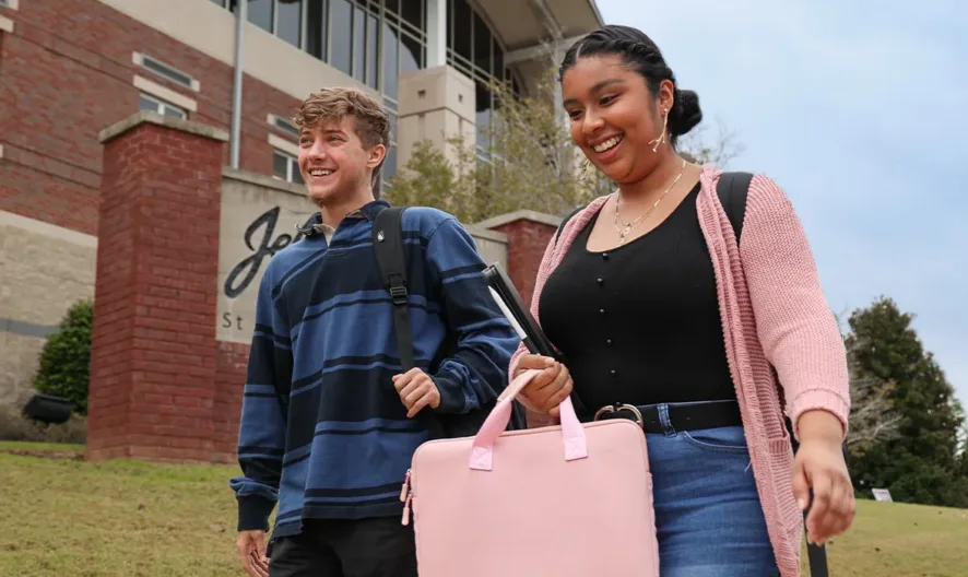Two smiling students walk outside a campus building. The young woman carries a large pink bag while the young man wears a striped shirt and backpack. The building and greenery are visible in the background.