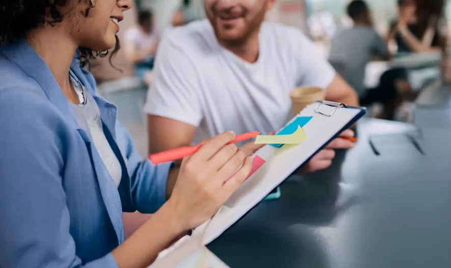 A woman and a man sit together at a table, discussing colorful sticky notes on a clipboard. The woman gestures with a red pen while other people work in the background in a casual, collaborative setting.