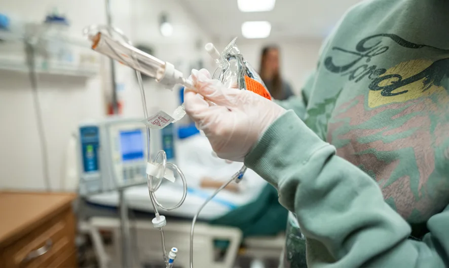 A person wearing gloves and a green Ford sweatshirt prepares an IV drip in a hospital room, with medical equipment and a patient bed visible in the background.