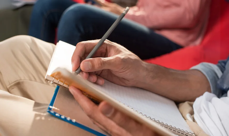 A person sits with a notebook on their lap, writing with a pen. They are wearing beige pants and a blue shirt, and another person is sitting beside them in the background.