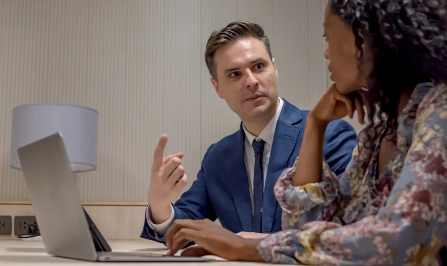 A man in a blue suit and a woman in a floral blouse sit at a table with a laptop, engaged in a serious conversation in a modern office setting.
