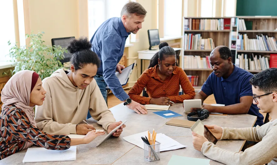 A group of six adults of diverse backgrounds sit and stand around a table in a library, engaging in discussion and looking at electronic devices and notebooks. Bookshelves and a plant are visible in the background.