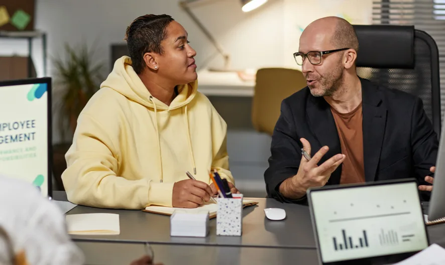 Two people sit at a desk, having a discussion and taking notes, while another person faces away. Computer monitors display graphs and Employee Engagement. The setting appears to be a modern office.