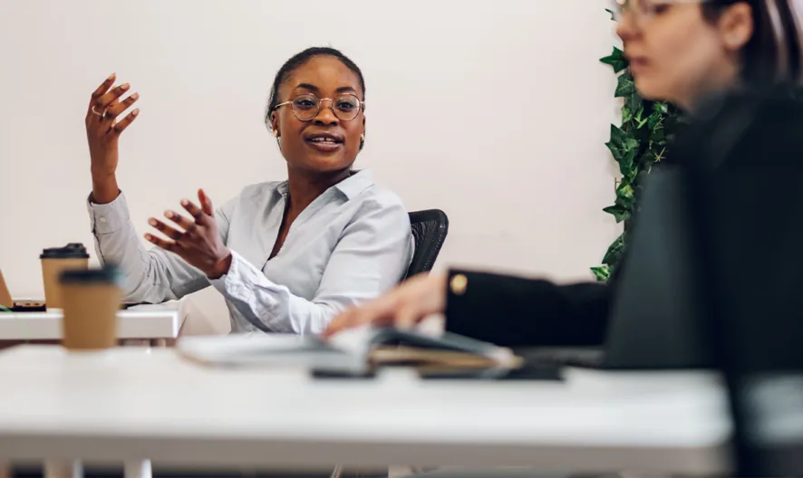 Two people sit at desks in an office. One person, wearing glasses and a light shirt, gestures while speaking to the other, who is blurred in the foreground. Laptops and a coffee cup are on the desks.