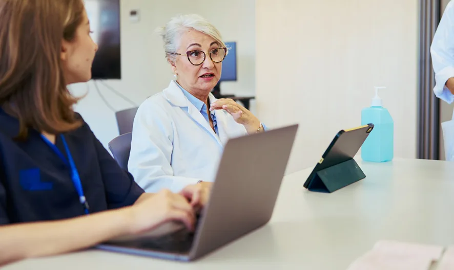 Three medical professionals, two seated and one standing, discuss in a modern office. One uses a laptop, another gestures while listening, and the third holds a clipboard. A tablet and sanitizer bottle are on the table.