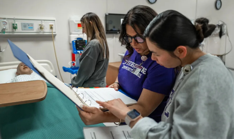 Two women review documents in a binder beside a hospital bed, while another person stands nearby and a patient lies in bed in the background. Medical equipment and a plant are also visible in the room.