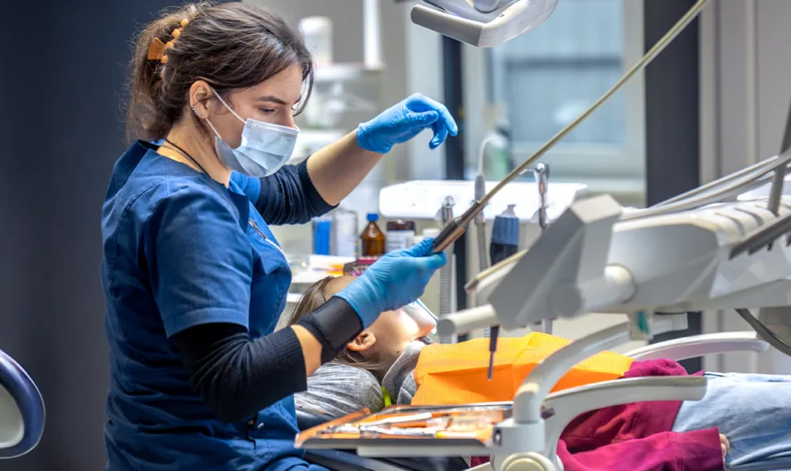 A dentist wearing a mask and gloves works on a young patient lying in a dental chair. The child is covered with an orange bib, and dental equipment surrounds them in a brightly lit clinic.