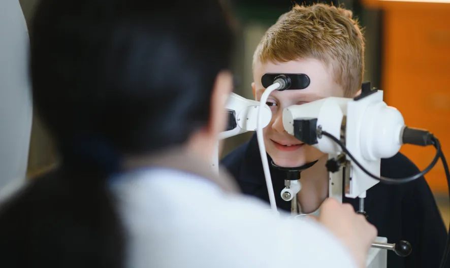 A child is having an eye exam with a slit lamp while a healthcare professional supervises, focusing on the childs eyes in a clinical setting.