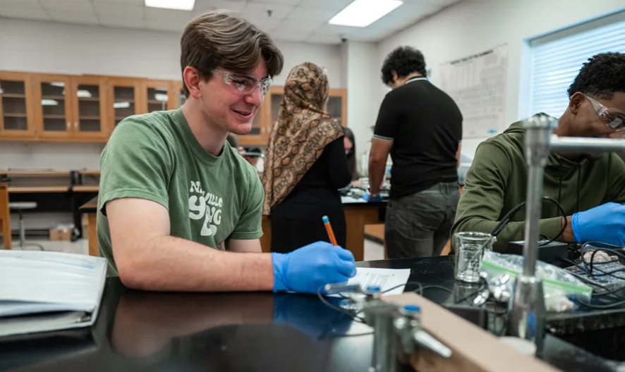 Two students wearing gloves and safety goggles work together at a lab table, smiling and writing notes. Other students are working in the background in a classroom with scientific equipment.