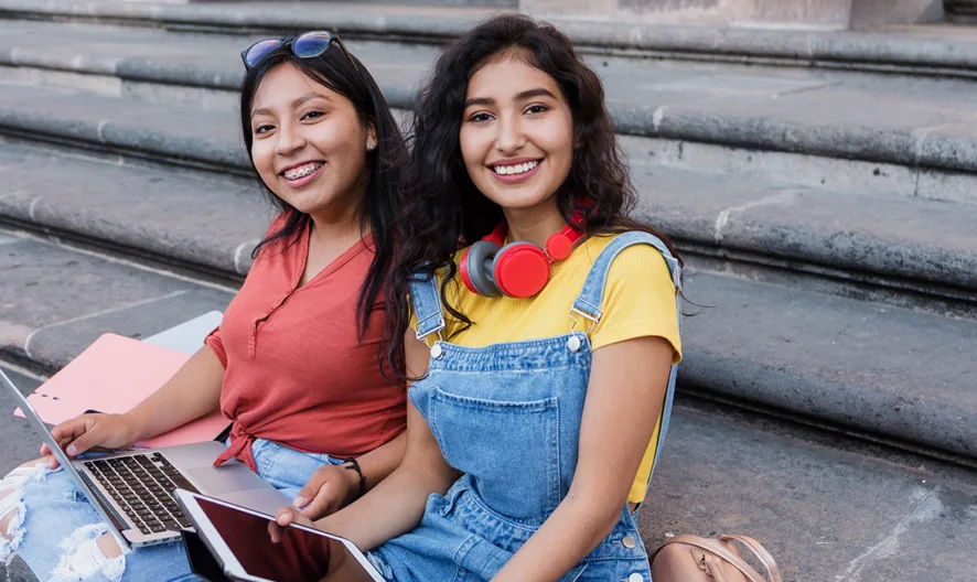 Two young women sit on outdoor steps smiling, with a laptop, notebook, and tablet in front of them. One wears sunglasses and holds a pink folder; the other wears red headphones and denim overalls.