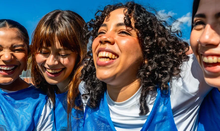 Four young women in blue sports jerseys smile and laugh together outdoors, with their arms around each other, enjoying a sunny day.