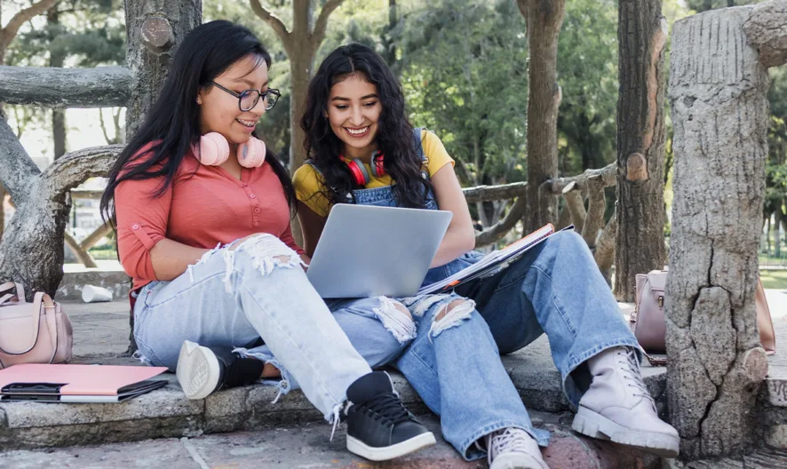 Two young women sit outdoors on stone steps, smiling and looking at a laptop. They are surrounded by notebooks, backpacks, and trees, suggesting they are studying together in a park or campus setting.