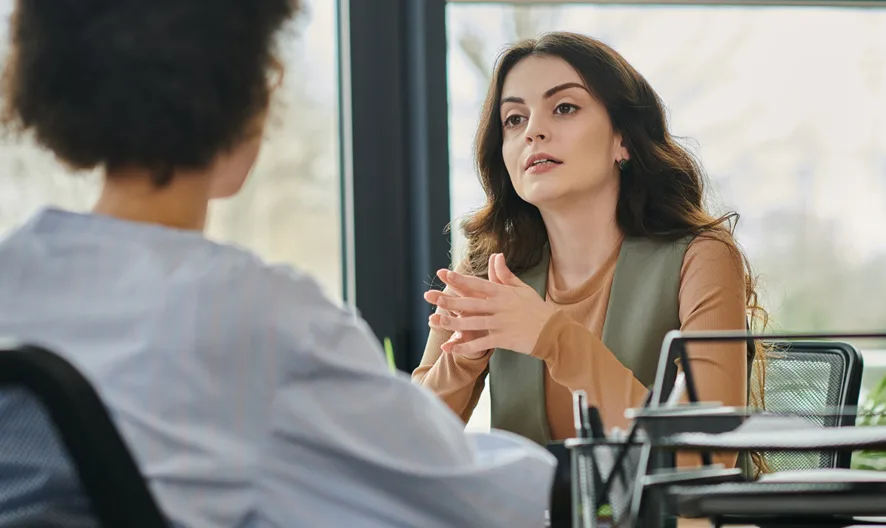 Two women sit across from each other at a desk in an office setting, engaged in a serious conversation. One woman is speaking with her hands clasped, while the other listens with her back to the camera.