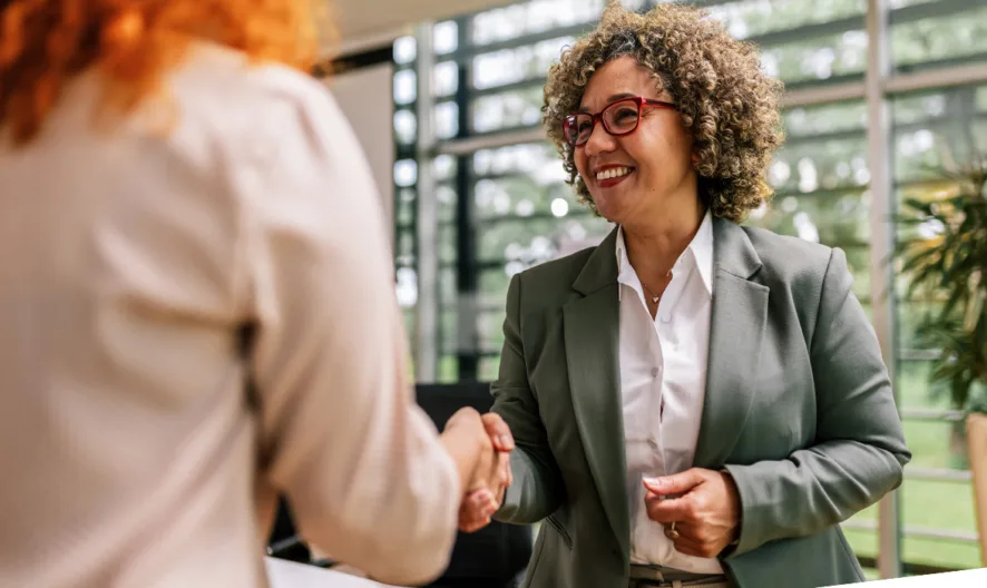 A woman with curly hair and glasses, dressed in a blazer, smiles and shakes hands with another person in a bright, modern office with large windows and plants in the background.