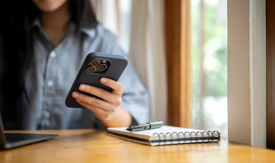 A person sits at a wooden table using a smartphone, with a laptop open in front of them and a spiral notebook with a pen nearby. Sunlight comes through large windows, creating a bright, airy workspace.