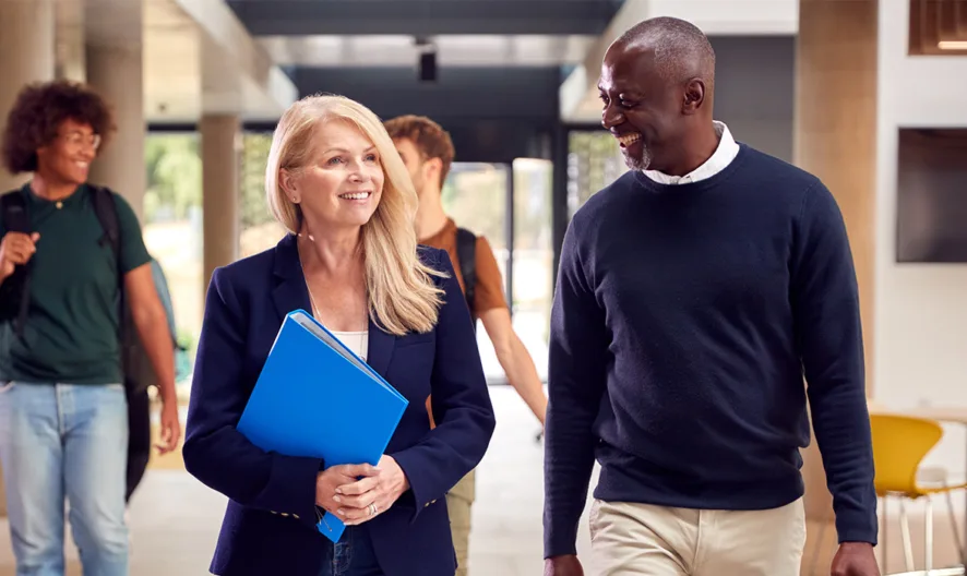 Two adults, a woman holding blue folders and a man in a dark sweater, walk and talk in a modern building hallway while two young people walk in the background.