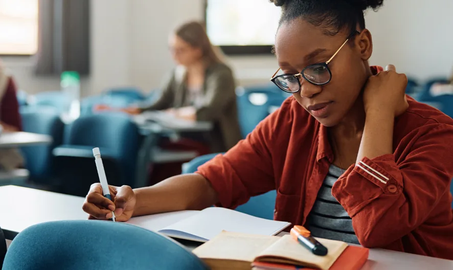 A student wearing glasses and a red shirt writes in a notebook in a classroom, surrounded by other students who are also studying or writing at their desks.