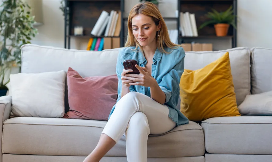 A woman sits on a gray couch with colorful pillows, looking at her smartphone. She is smiling and indoors, with bookshelves and plants in the background.
