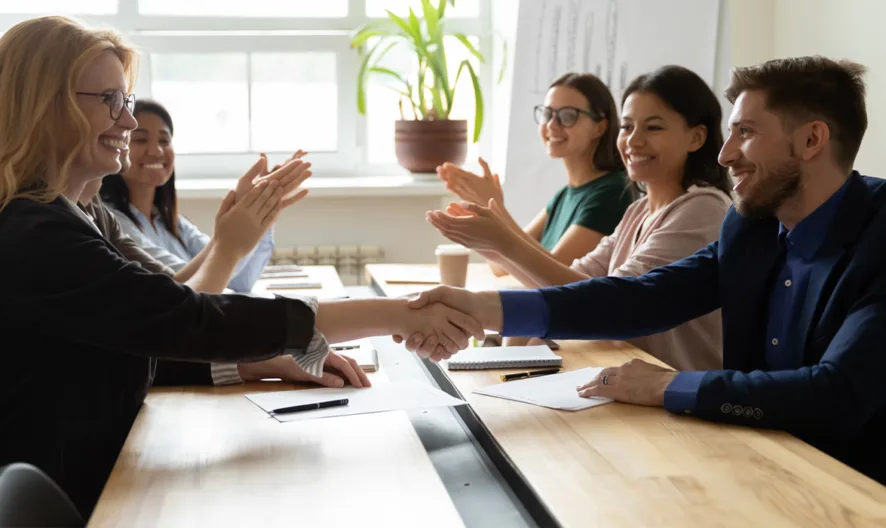 Five people sit at a conference table. A woman and a man are shaking hands and smiling, while three others sit nearby, watching and clapping. Sunlight streams through a window with a potted plant in the background.