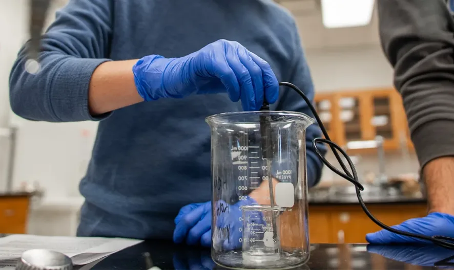 Two people wearing blue gloves work in a lab, one holding a sensor or probe in a large glass beaker on a black lab table, with shelves and cabinets visible in the background.
