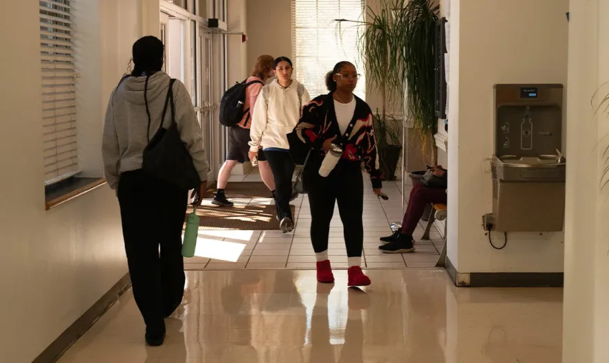 Students walk and talk in a well-lit hallway with large windows, a water fountain, and a potted plant. Some students are carrying backpacks, and natural light fills the space.