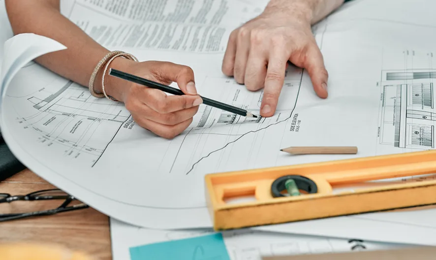Two people working on architectural blueprints at a desk, one person drawing with a pencil while the other points at the plans; a yellow level, glasses, and other drawing tools are on the table.