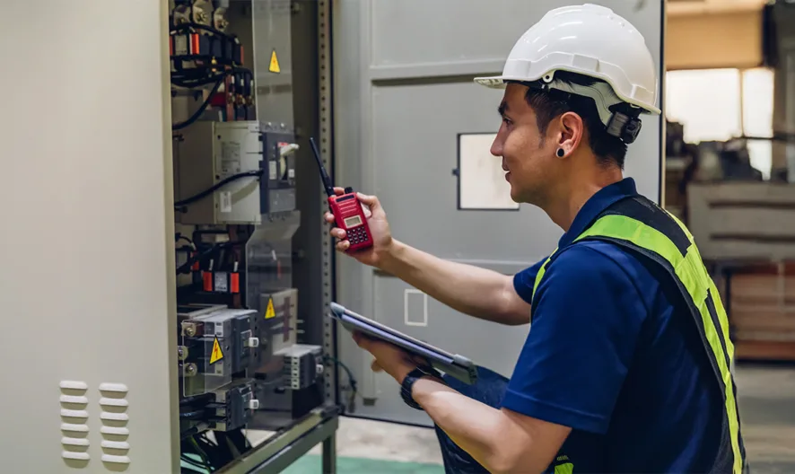 A male worker in a white hard hat and safety vest inspects electrical equipment inside an open control panel, holding a digital device in one hand and a clipboard in the other, in an industrial setting.