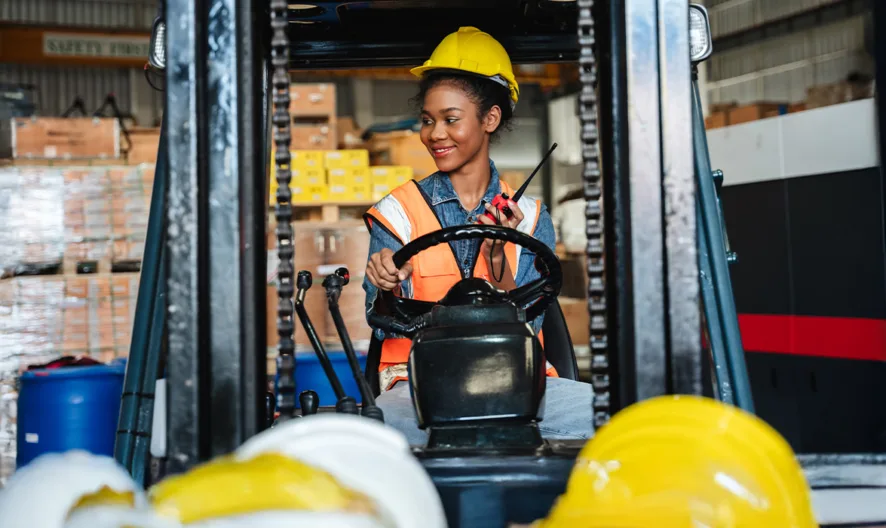 A woman wearing a safety vest and yellow hard hat is smiling while operating a forklift in a warehouse filled with stacked boxes and safety helmets.