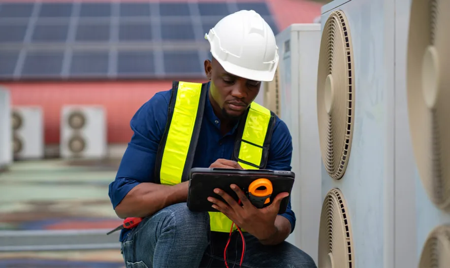 A worker wearing a white hard hat and yellow safety vest uses a tablet while inspecting large industrial HVAC units on a rooftop.