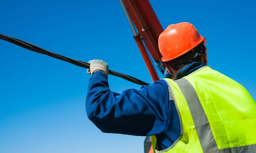 A worker in a neon safety vest and orange hard hat holds a thick black cable while looking upward, with a clear blue sky in the background.
