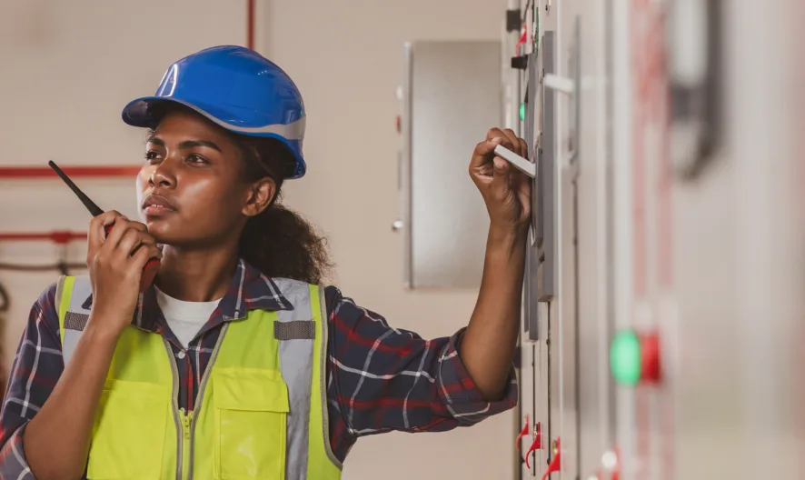 A woman wearing a blue hard hat, safety vest, and plaid shirt operates a control panel while speaking into a walkie-talkie in an industrial setting.