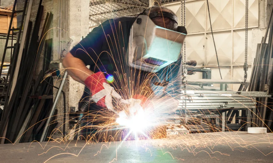 A person wearing protective gear and a welding helmet uses a welding torch, producing bright sparks, in an industrial workshop surrounded by metal structures and equipment.