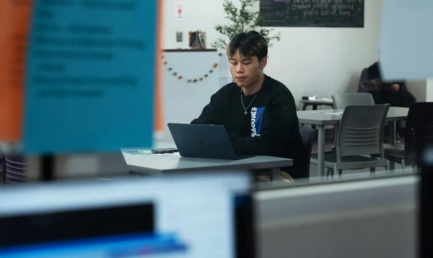 A person sits alone at a table in a modern, bright room, working on a laptop. Other people and colorful notes on glass are visible in the background, creating a focused, academic atmosphere.