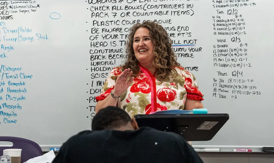 A smiling teacher stands at a podium in front of a whiteboard covered in handwritten notes, speaking to students seated at desks in a classroom.