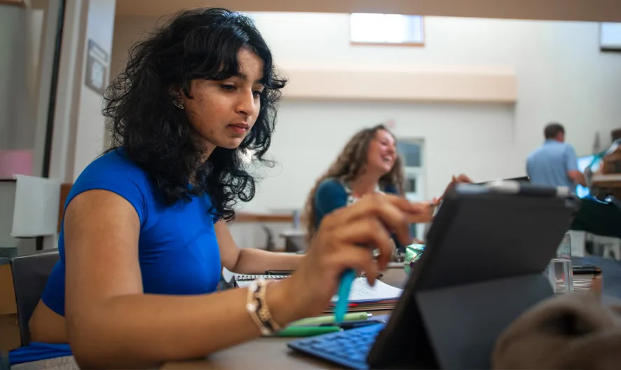 A woman with dark curly hair, wearing a blue top, sits at a desk using a tablet. Other people are working and talking in the background in a bright, modern office space.