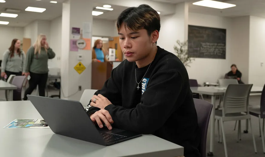 A student sits at a table in a bright, modern lounge area, working on a laptop. Several people are talking in the background near a doorway, and other students are seated throughout the room.