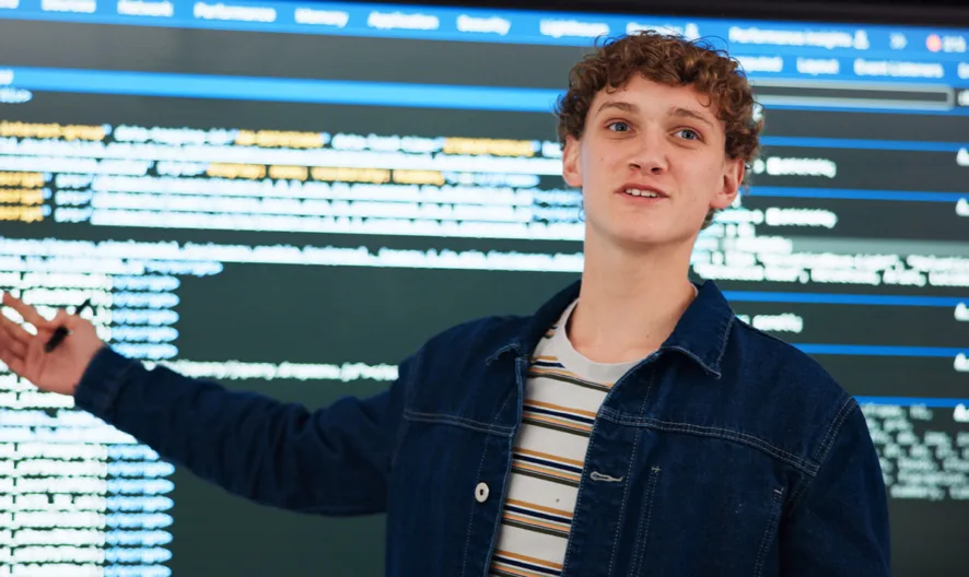 A young person with curly hair points to a large display screen filled with lines of code, appearing to explain or present something. The individual wears a striped shirt and denim jacket.