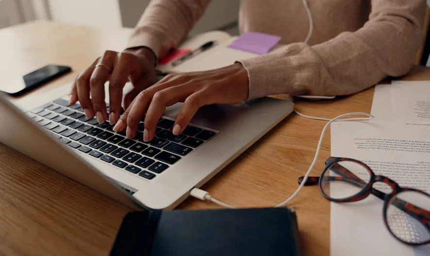 A person types on a laptop at a wooden desk, surrounded by documents, a pair of glasses, a notebook, and a smartphone. Earphones are plugged into the laptop. The setting appears to be a home or office workspace.