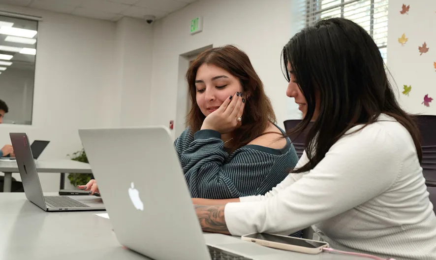 Two women sit at a table using laptops, collaborating and smiling in a brightly lit room with fall decorations on the wall. Other people work at separate tables in the background.