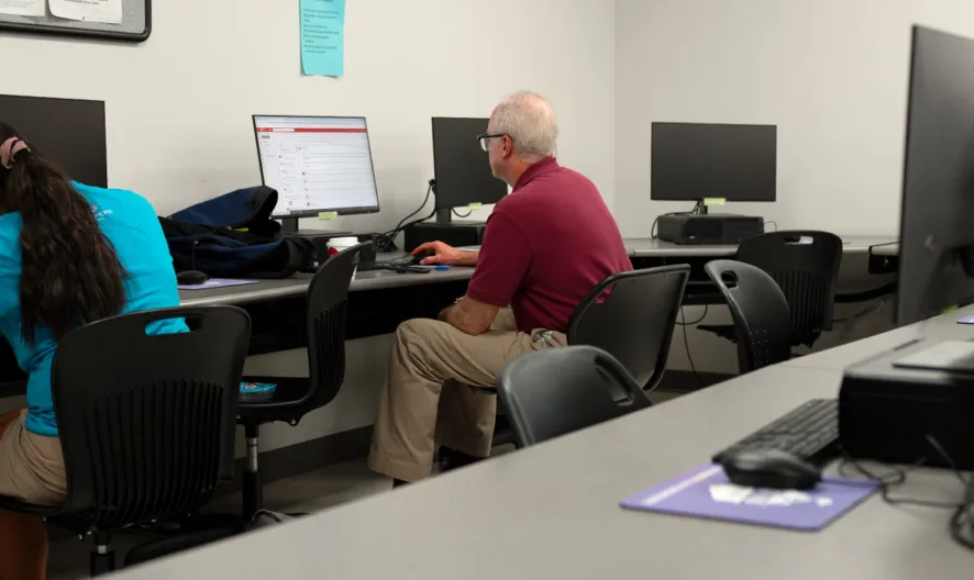 Two people sit at separate desks using computers in a classroom or computer lab setting. Several other computer workstations are visible but unoccupied. Papers and bags are on the desks.