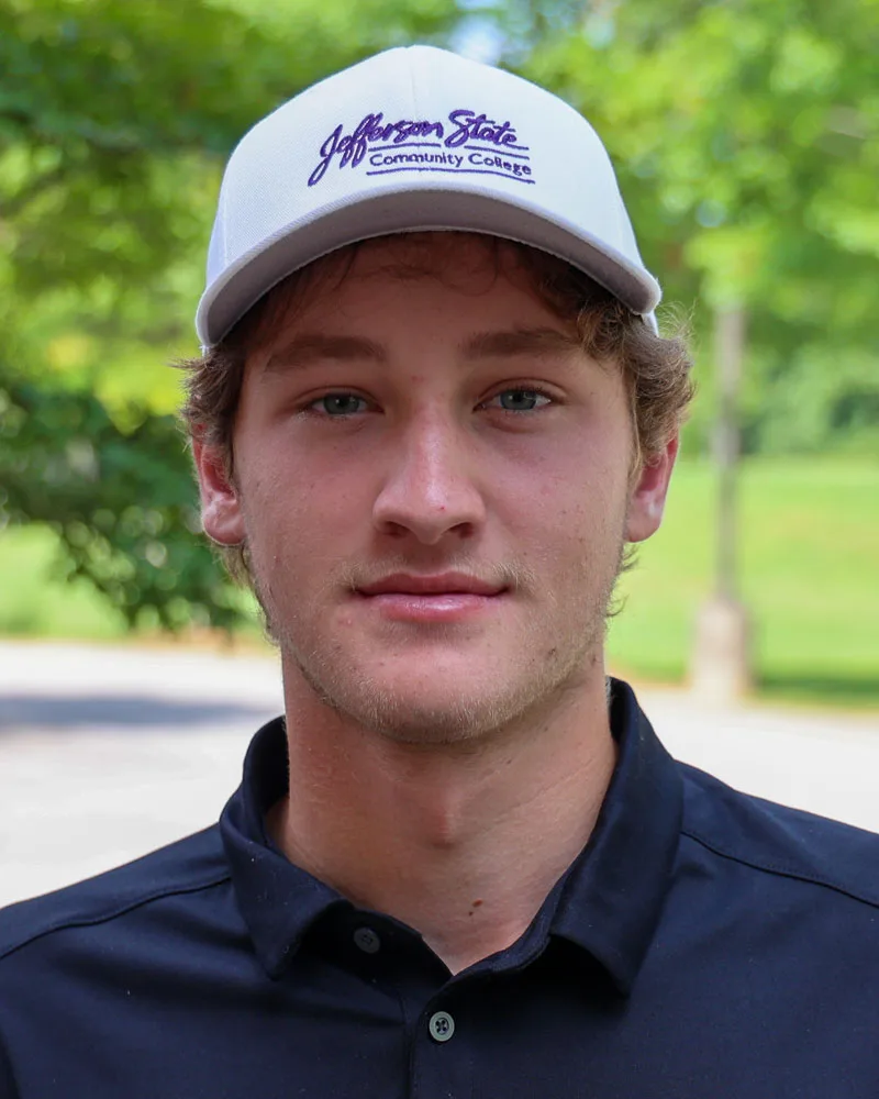 A young man with light brown hair, wearing a white Jefferson State Community College cap and a dark collared shirt, stands outdoors with greenery and a blurred path in the background.