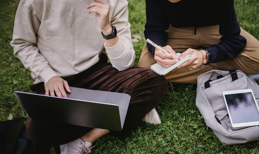 Two people sit on grass. One uses a laptop while the other writes in a notebook. A backpack and a tablet are placed nearby. Both appear focused, working or studying together outdoors.