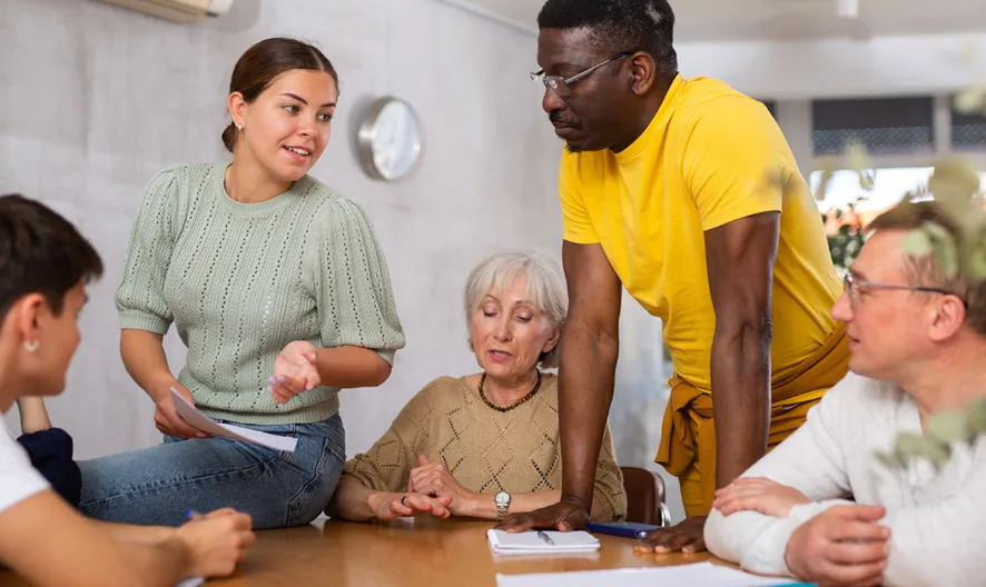 Five people of diverse ages and backgrounds are gathered around a table, engaging in a discussion. One woman speaks while others listen attentively, creating a collaborative and thoughtful atmosphere.