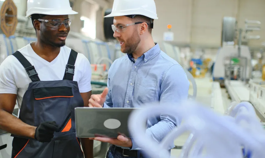 Two men wearing safety helmets and glasses stand in a factory, one in work overalls and gloves, the other in a shirt holding a laptop. They are having a discussion surrounded by industrial equipment.