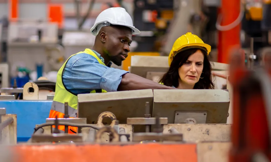 Two factory workers, one man and one woman, both wearing safety helmets and high-visibility vests, examine industrial equipment together in a busy manufacturing facility.