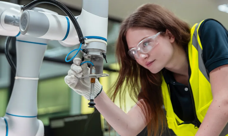 A woman wearing safety goggles, a yellow safety vest, and a white glove operates or inspects a robotic arm in a modern industrial or laboratory setting.