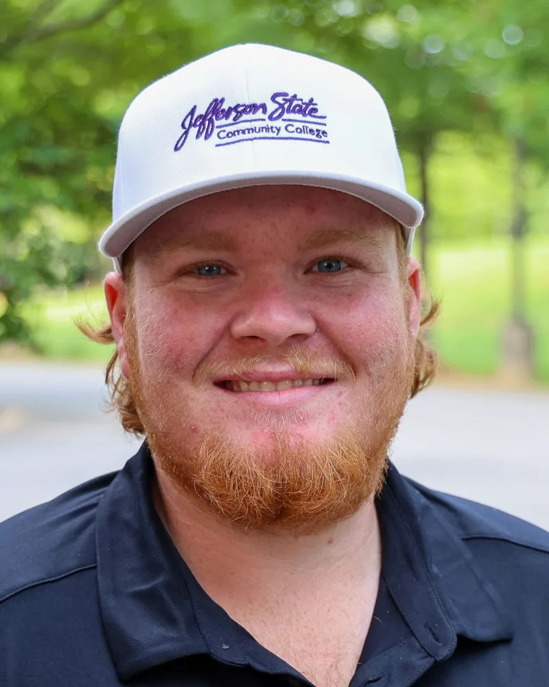 A young man with a red beard smiles outdoors. He wears a white Jefferson State Community College cap and a black collared shirt, with green trees blurred in the background.
