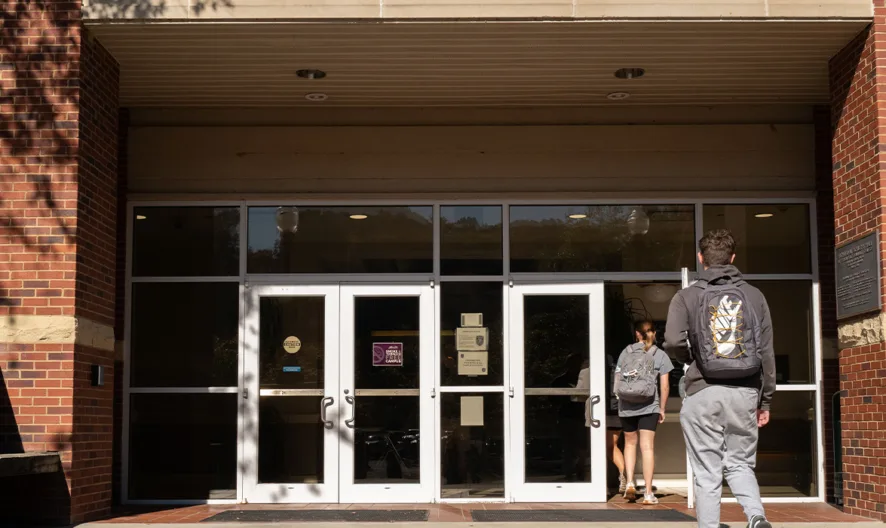 Two students with backpacks walk toward the glass entrance doors of a red brick building on a sunny day.