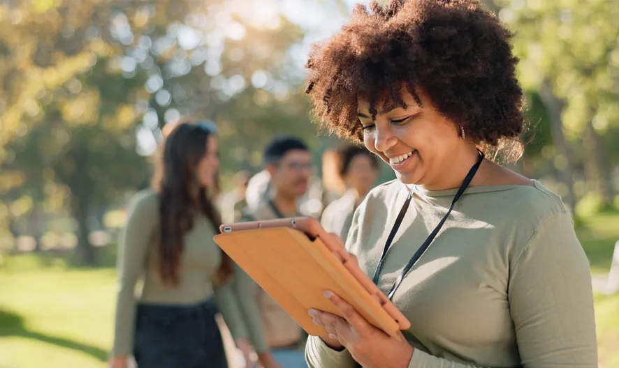 A smiling woman stands in a sunlit park, holding and looking at a tablet. She wears a green top and lanyard, with a group of people and trees blurred in the background.