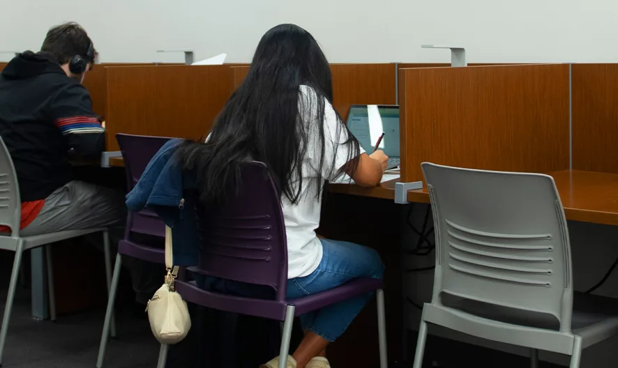 Several students sit at individual wooden study cubicles, working on laptops and writing. The setting appears to be a quiet study area in a library or learning center.